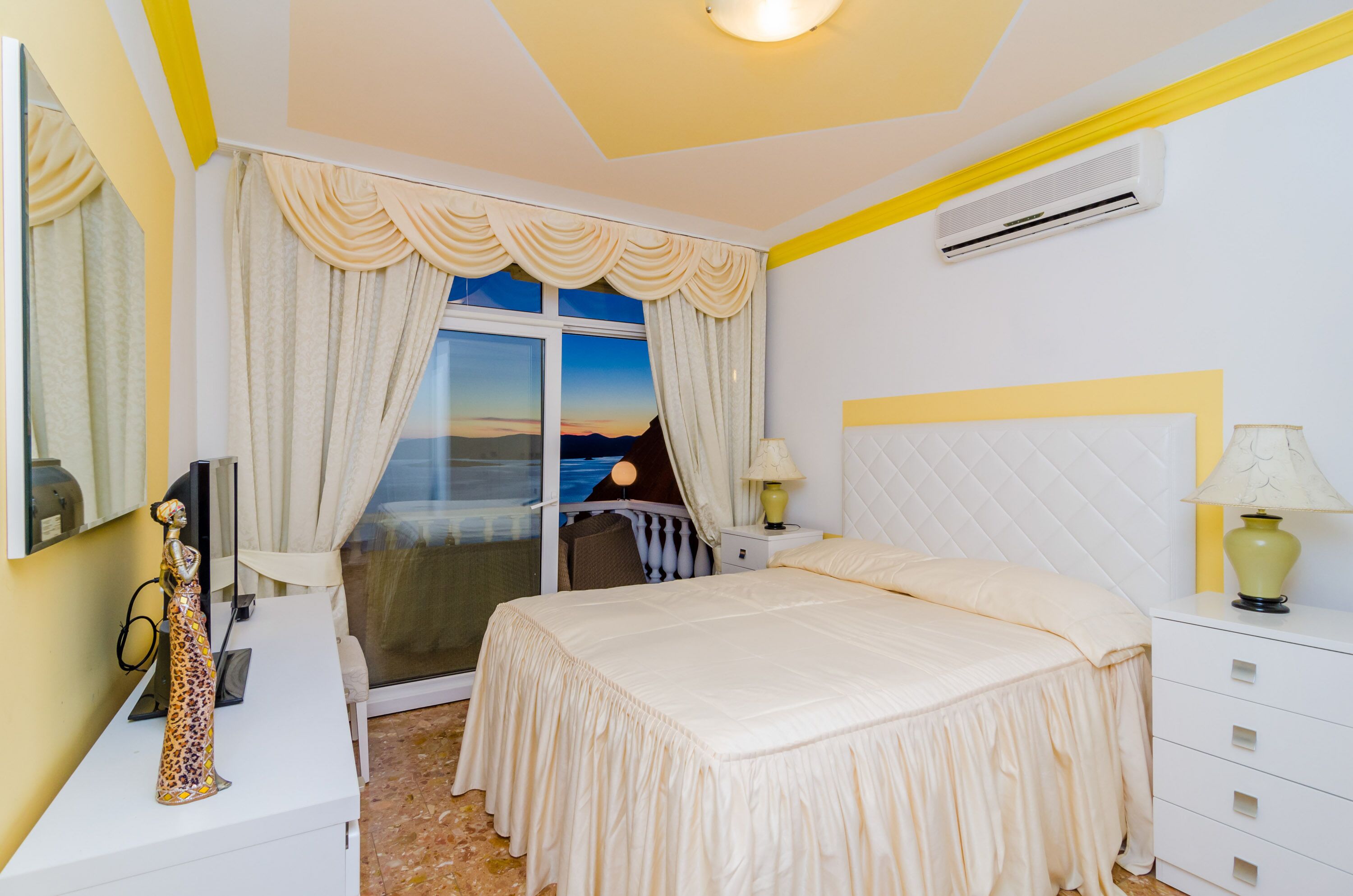 A hotel room with a bed, bedside tables, a lamp, a window with curtains, and a view of the sunset.