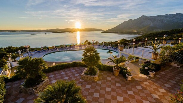 Una piscina de resort con suelo a cuadros, rodeada de sillas de descanso y plantas en macetas, con vistas a un cuerpo de agua y montañas.
