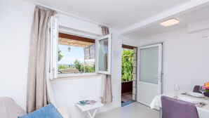 Quarto com mesa de jantar posta para dois, uma pequena mesa branca com revistas, janela com cortinas e vista para o verde lá fora.