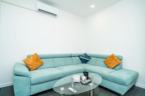 En teal farvet sofa med to orange og en blå pude, et glas sofabord med en fjernbetjening, og en hvid klimaanlæg på væggen.