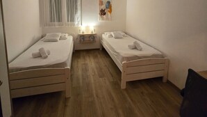 Une petite chambre propre, avec deux lits simples, une table de chevet avec une lampe et une fenêtre avec des rideaux.