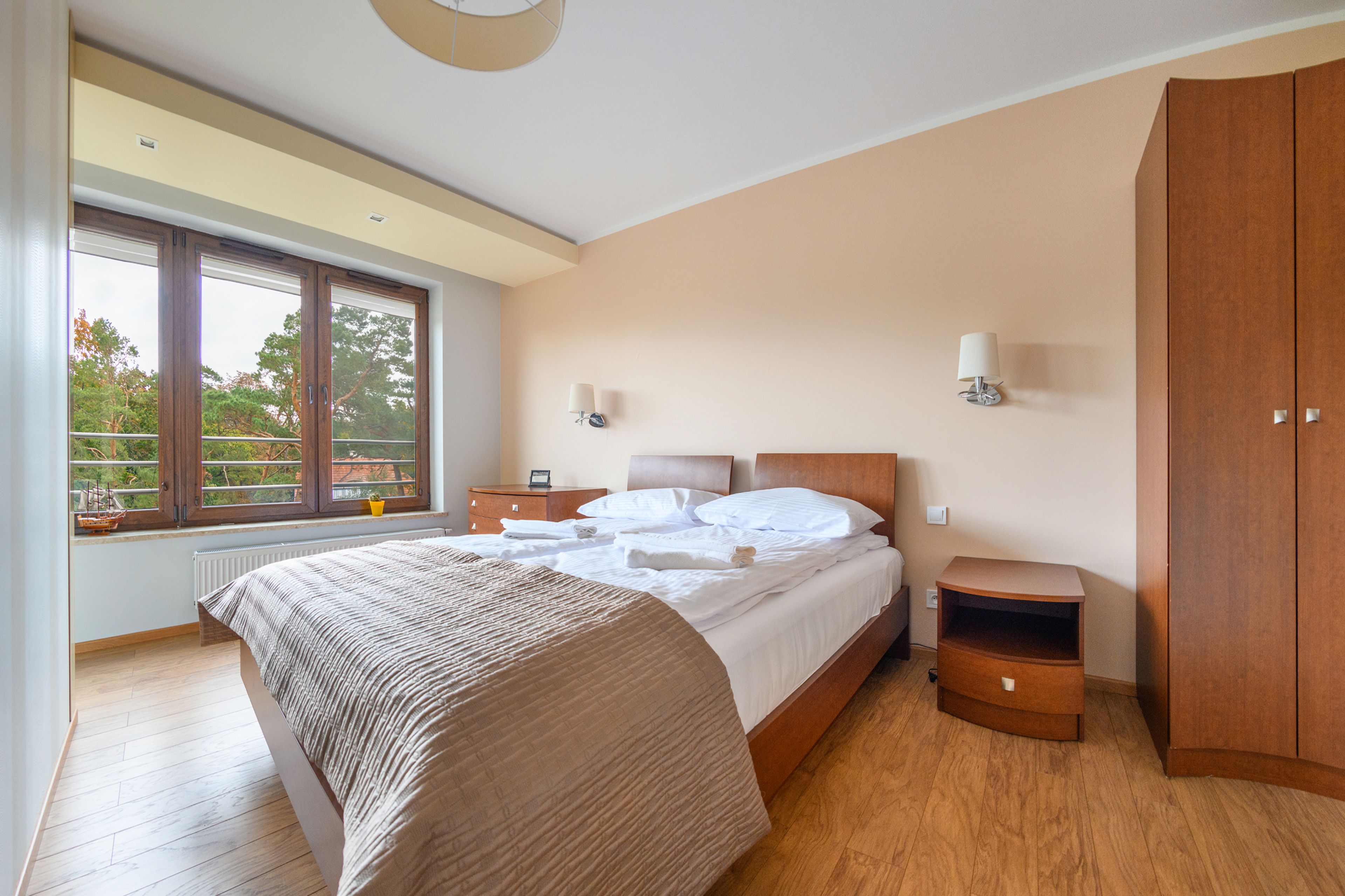 A bedroom with a large bed, wooden wardrobe, bedside tables, and a window with a view of trees.