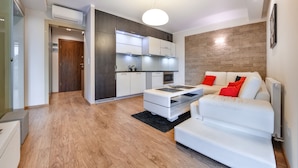 Una sala de estar moderna con un sofá blanco, una mesa de centro de vidrio y una cocina con armarios blancos y una pared de acento de madera.