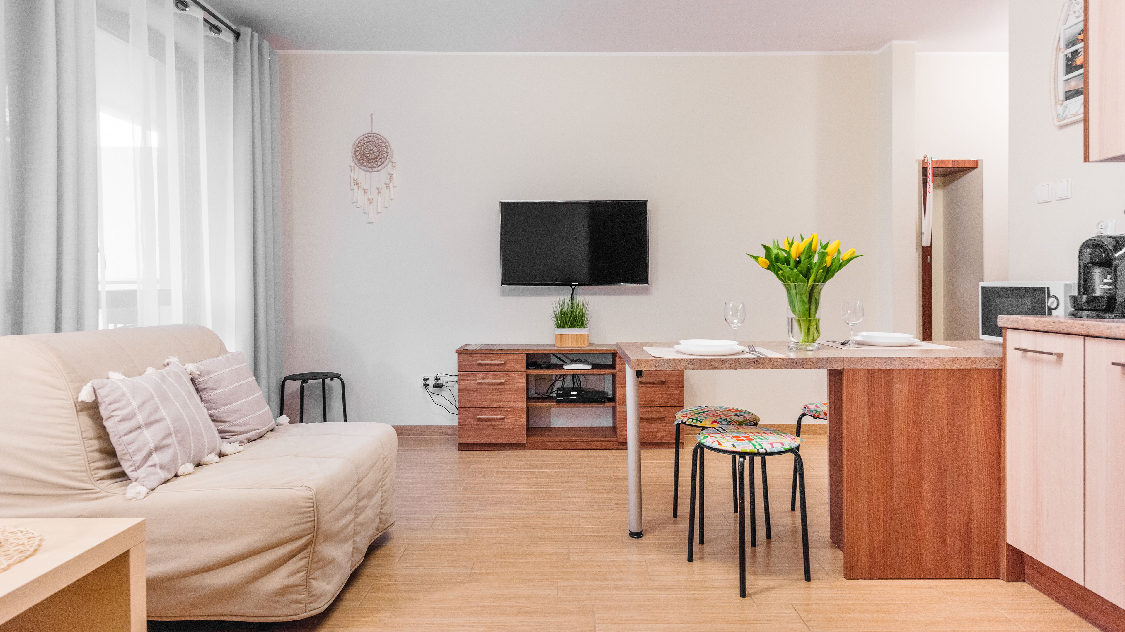 A modern, well-lit living room with a beige sofa, a small dining table with two chairs, a TV mounted on the wall, and a kitchen area with a coffee machine.