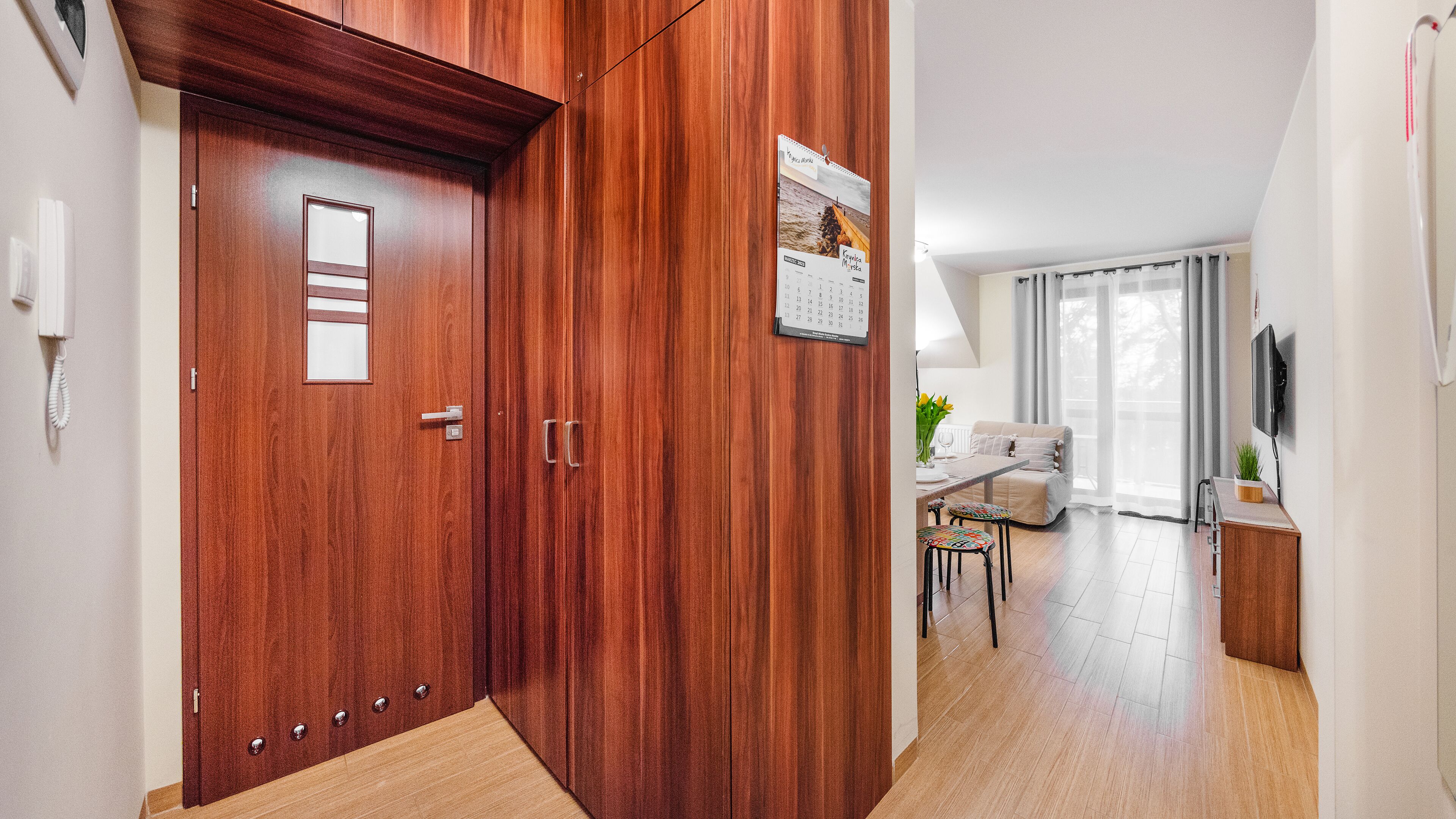 Una puerta de madera con un calendario, una mesa pequeña con un jarrón y un sofá en una habitación luminosa con un televisor.