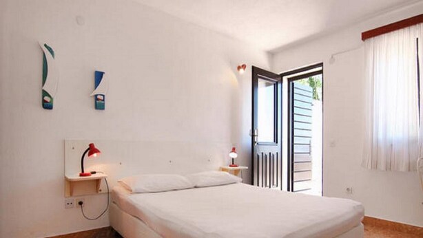 A bedroom with a bed, a nightstand, a red lamp, a white wall, a window with curtains, and a door leading outside.
