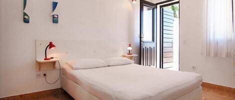 A bedroom with a bed, a nightstand, a red lamp, a white wall, a window with curtains, and a door leading outside.