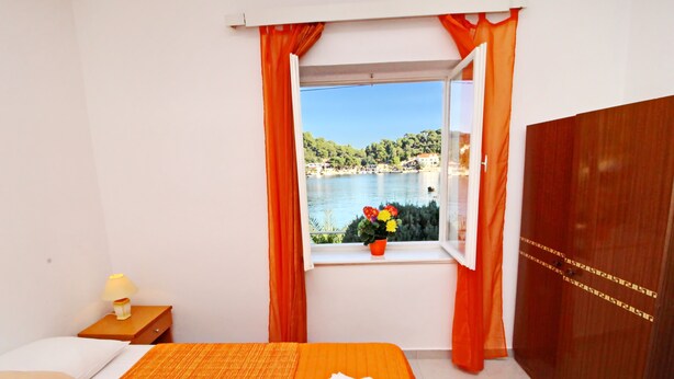 Habitación de hotel con cama, mesita de noche, ventana con cortinas y vistas a un lago y edificios.
