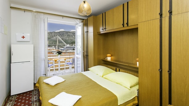A hotel room with a bed, a small table, a wardrobe, a bedside table with a lamp, a window with a view of a bridge and boats, and a door with a red cross sign. - Guest House Bridge View - Double Room with Balcony and Sea View (Dubrovnik)