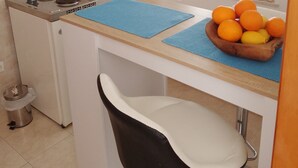Une cuisine compacte équipée d’un évier, d’une cuisinière et d’un petit coin repas avec une chaise et un panier de fruits.