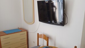 Une petite pièce comprenant un bureau en bois, une chaise, un téléviseur fixé au mur, une commode et un tableau représentant un paysage marin.