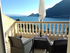 Un balcon agrémenté de meubles en osier, une table avec une bouteille et des verres, et une vue sur la mer et les montagnes.