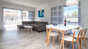 A modern living room with a gray sectional sofa, a round coffee table, and a dining table with chairs. There is a large window with sheer curtains and a view of buildings outside. - Dva Galeba - One Bedroom Apartment with Garden Terrace (202) (Bol)