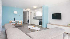 A modern living room with a sofa, a dining table with chairs, a kitchen area, and a TV mounted on the wall. - Dva Galeba - One Bedroom Apartment with Garden Terrace (202) (Bol)