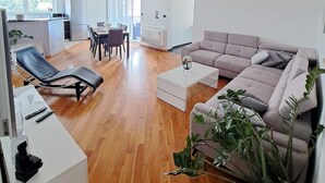 Un salon moderne avec un canapé, une table basse et un fauteuil. On aperçoit à l’arrière-plan un coin repas avec une table et des chaises.