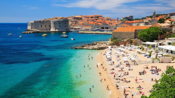 Une ville côtière dotée d’un fort historique, d’eaux turquoise cristallines et d’une plage de sable où l’on trouve des parasols et des personnes qui prennent des bains de soleil.