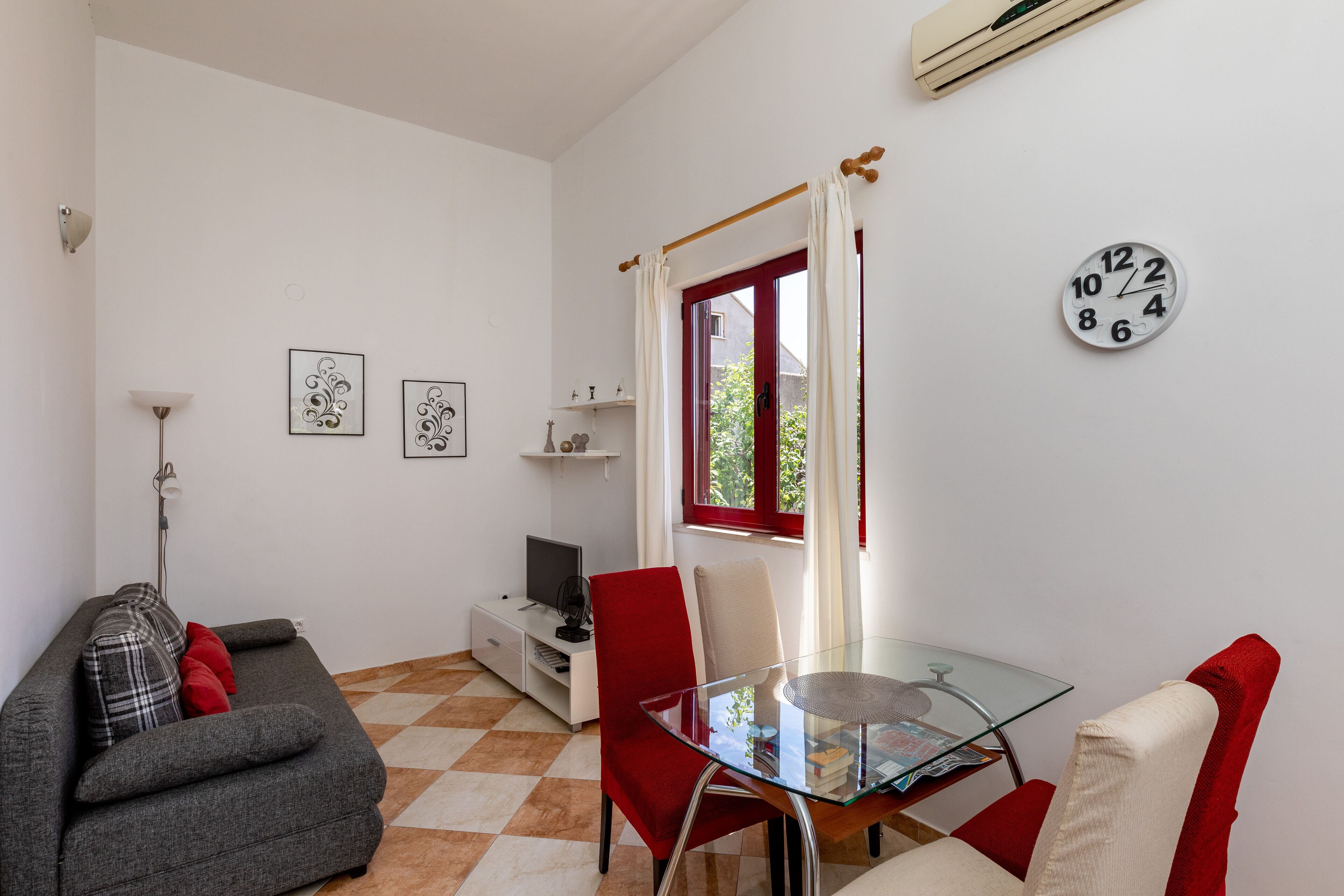 A modern living room with a glass dining table, red chairs, a gray sofa, and a red and white checkered floor.