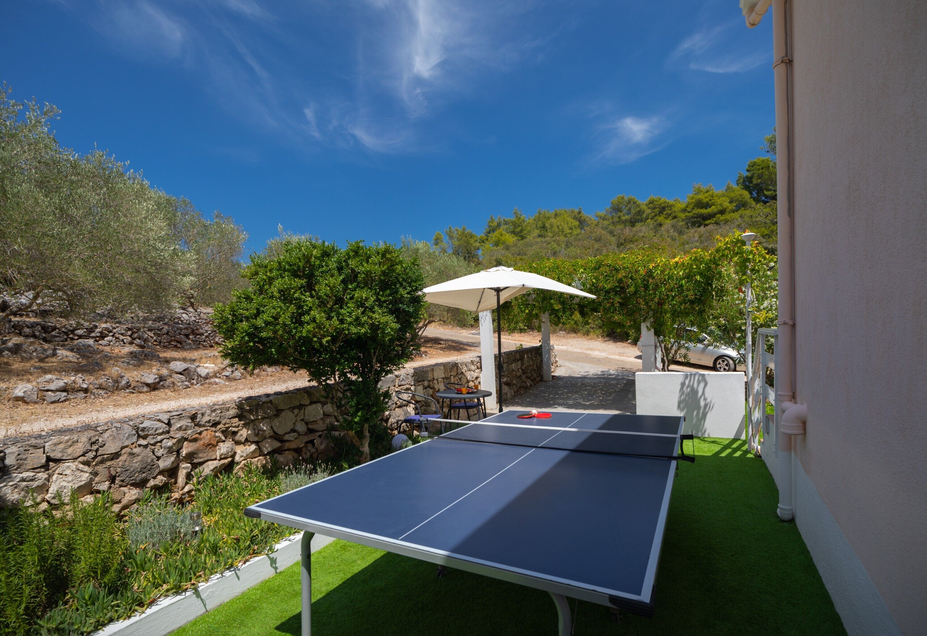 A ping pong table in a garden with a stone wall, a white gazebo, and a car in the background.