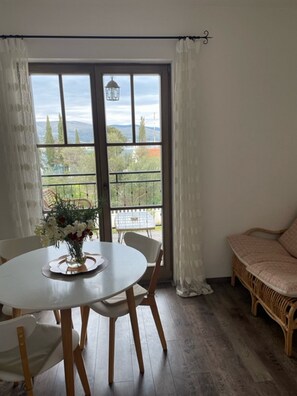 Un coin repas avec une table ronde, des chaises et un vase de fleurs. Il y a un balcon avec une balustrade et une vue sur la verdure et les bâtiments.
