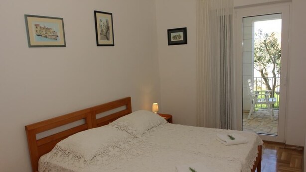 A bedroom with a wooden bed, white bedding, and a nightstand with a lamp. There are framed pictures on the wall and a view of the outdoors through a glass door. - Apartments Villa Muškat - Comfort Two Bedroom Apartment with Terrace (Supetar)