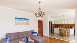 Un salon avec un canapé, une petite table et un lustre. On aperçoit la cuisine, avec une table à manger et des chaises.