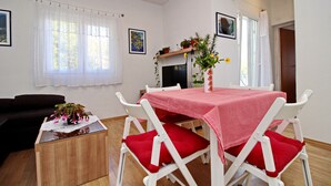 A dining area with a table covered by a red tablecloth, white chairs with red cushions, a wooden side table with a vase of flowers, and a television on a stand.