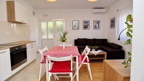 A modern kitchen with white cabinets, a built-in oven, and a wooden countertop. There is a dining area with a pink table and white chairs. A black sofa and a small wooden table are in the living area.