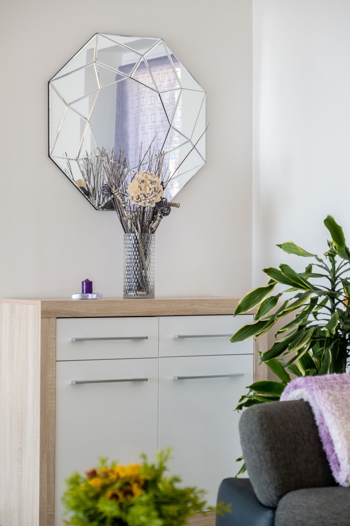 Et værelse med et geometrisk spejl, en vase med tørrede blomster, et skab med skuffer og en plante.