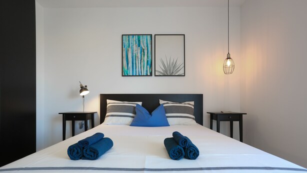 Una cama bien hecha con ropa de cama azul y blanca, dos mesitas de noche con lámparas y dos obras de arte enmarcadas en la pared.