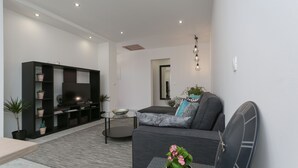Una sala de estar moderna con un sofá, una mesa de centro y un televisor sobre un mueble. Se observa una estantería con plantas y una pequeña mesa con un cuenco.