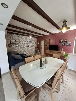 Un'area pranzo con un tavolo bianco, sedie di legno, una parete in pietra e una televisione sullo sfondo.