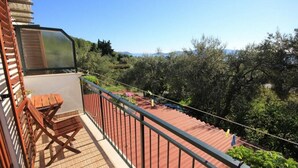 Un balcon avec un banc en bois, une rambarde en métal et une vue sur des oliviers et des collines.