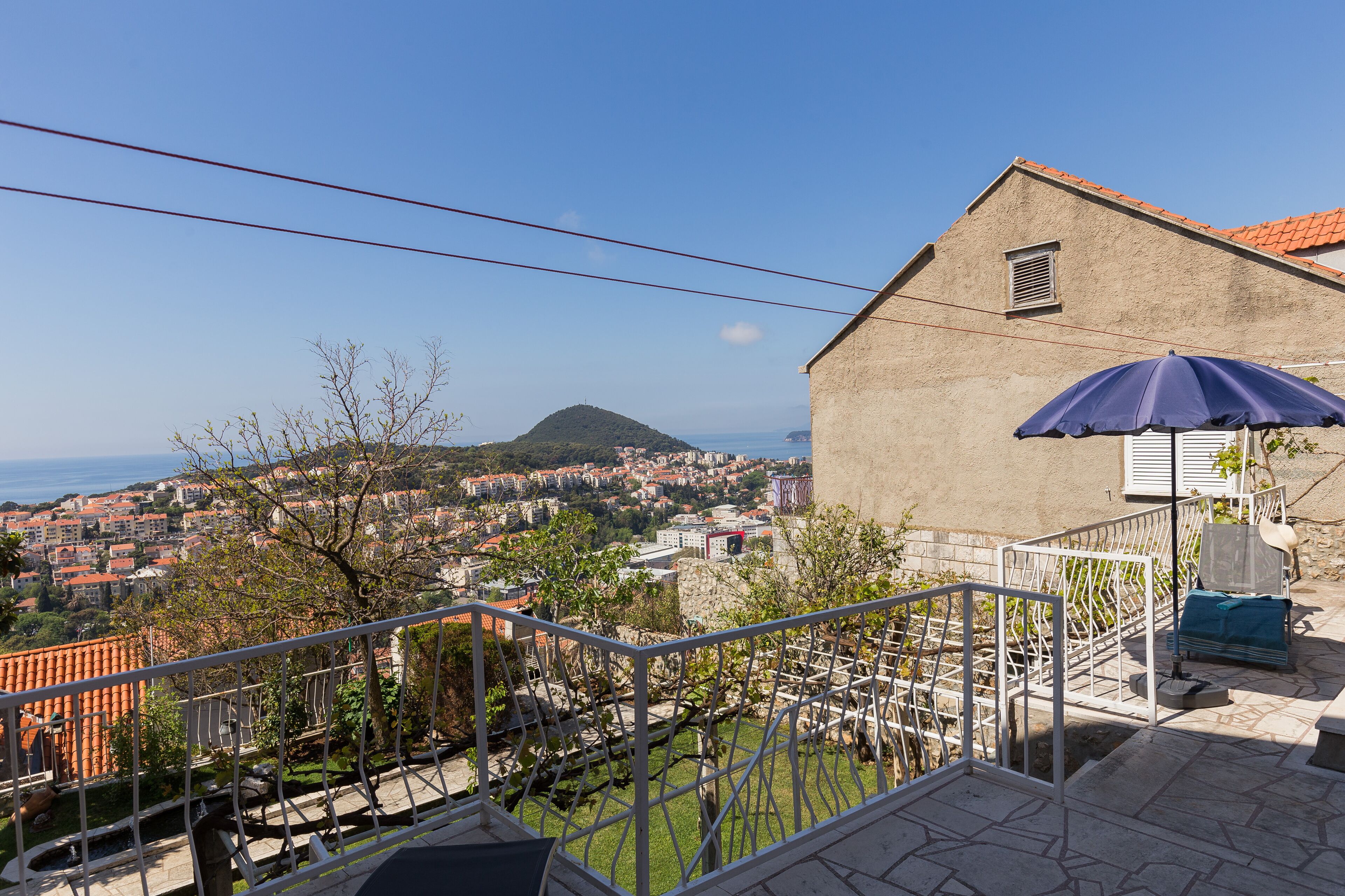 A building with a red-tiled roof, a balcony with a metal railing, and a view of a town and a hill in the distance.