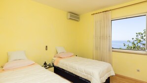 A room with two beds, a window with a view of the sea, and an air conditioning unit on the wall.