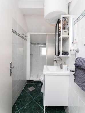 A bathroom with a white sink, mirror, and toilet. There is a shower area with a glass door and a small window. The floor is covered with green marble tiles.