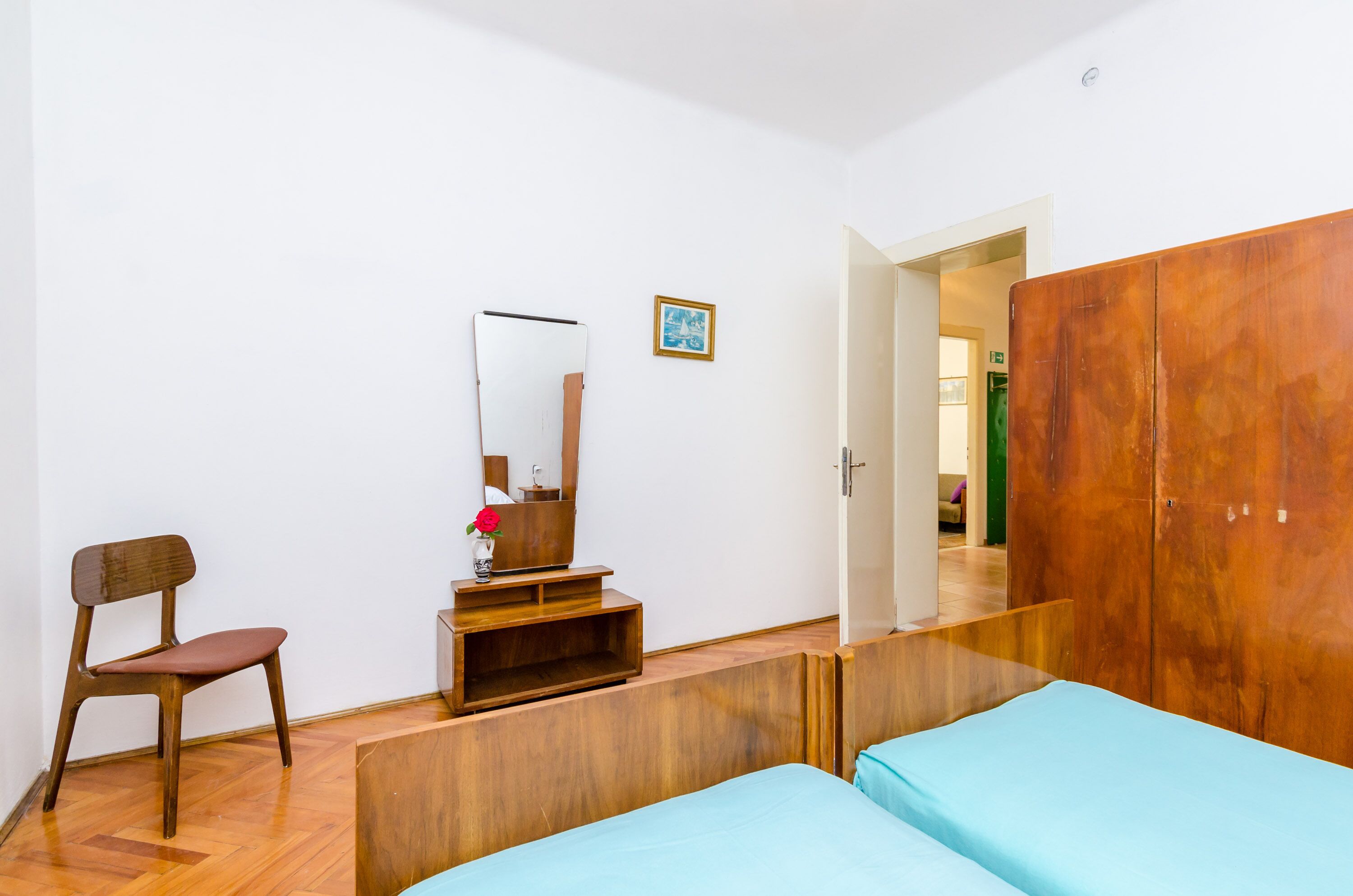 Un dormitorio con un armario de madera, una silla de madera, una cama con colcha azul, una mesita de madera con un jarrón y una flor roja, y un espejo en la pared.