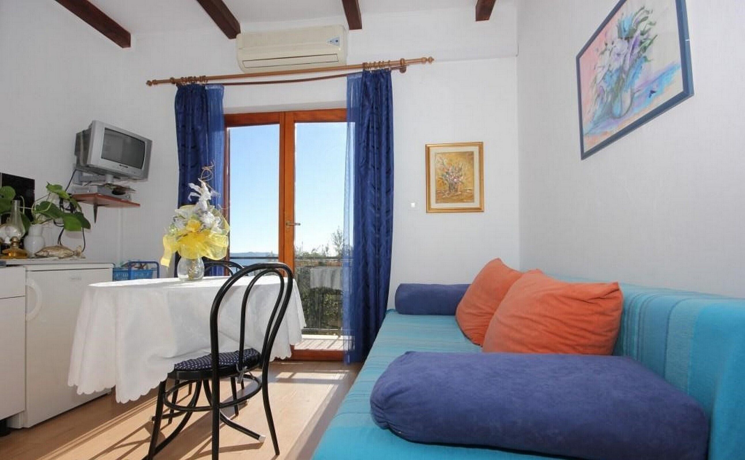 Une pièce avec un canapé bleu, une petite table avec une chaise, un téléviseur et une fenêtre avec des rideaux.