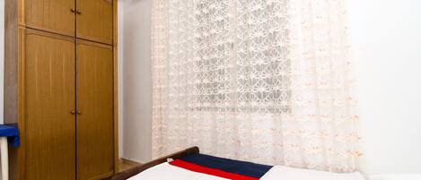 Een slaapkamer met een houten garderobe, een bed met een wit en rode deken en een doorschijnend gordijn met een bloemenprint.