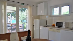 Eine Küche mit weißen Schränken, einer Mikrowelle und einem Kühlschrank. Ein Esstisch mit Stühlen und eine Weinflasche stehen bereit.