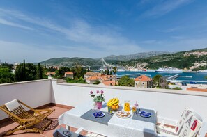 Una terrazza con un tavolo apparecchiato per mangiare, affacciata su una cittadina costiera con un ponte e montagne sullo sfondo.