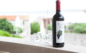 A bottle of Plavac Mali wine and a broken glass on a balcony railing with a view of buildings and greenery in the background.