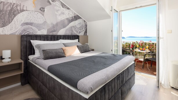 Une chambre moderne avec un grand lit, une vue sur la mer grâce à une porte-fenêtre coulissante et un balcon doté d’un espace salon extérieur.