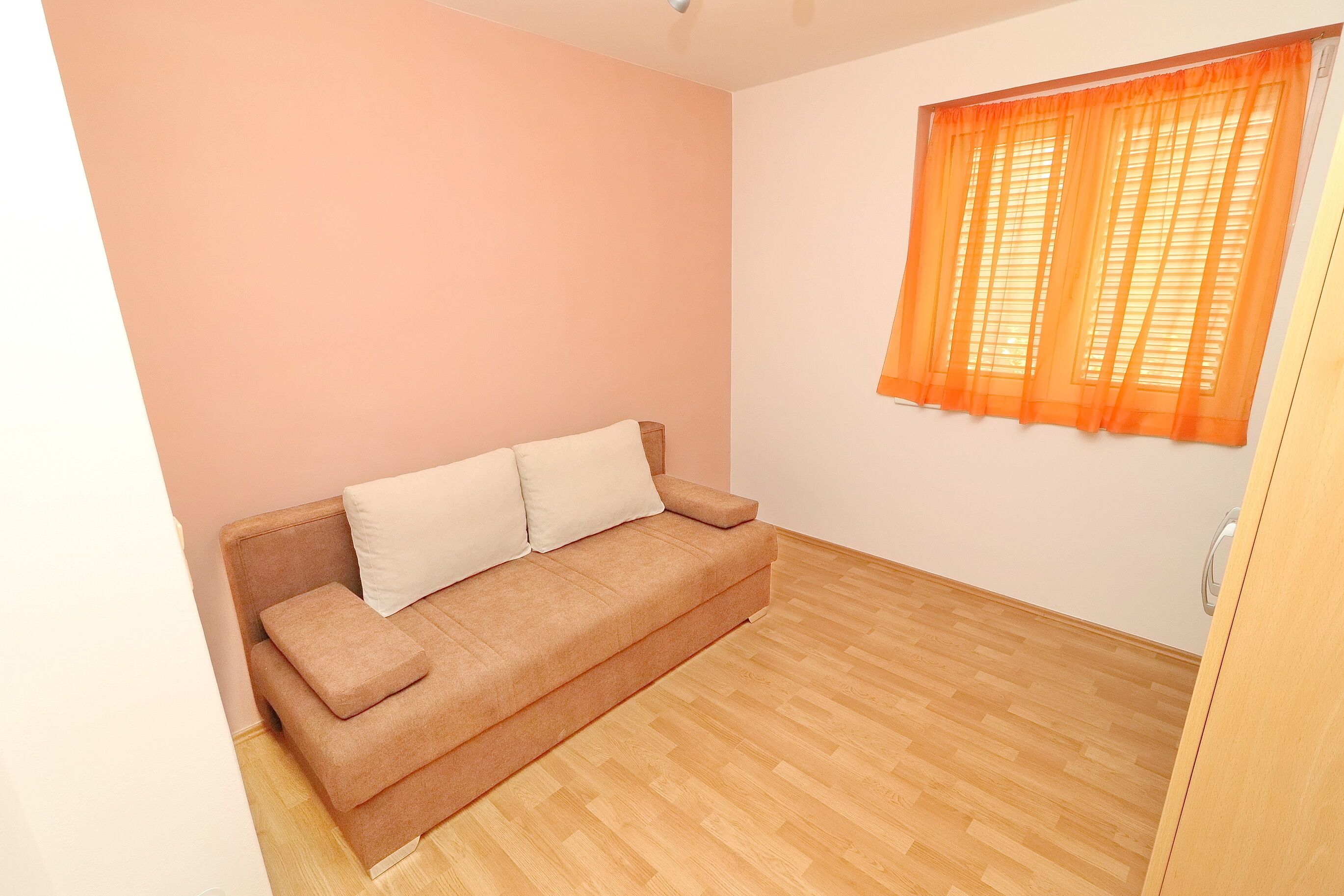 A room with a beige sofa, a wooden floor, and an orange window with sheer curtains.