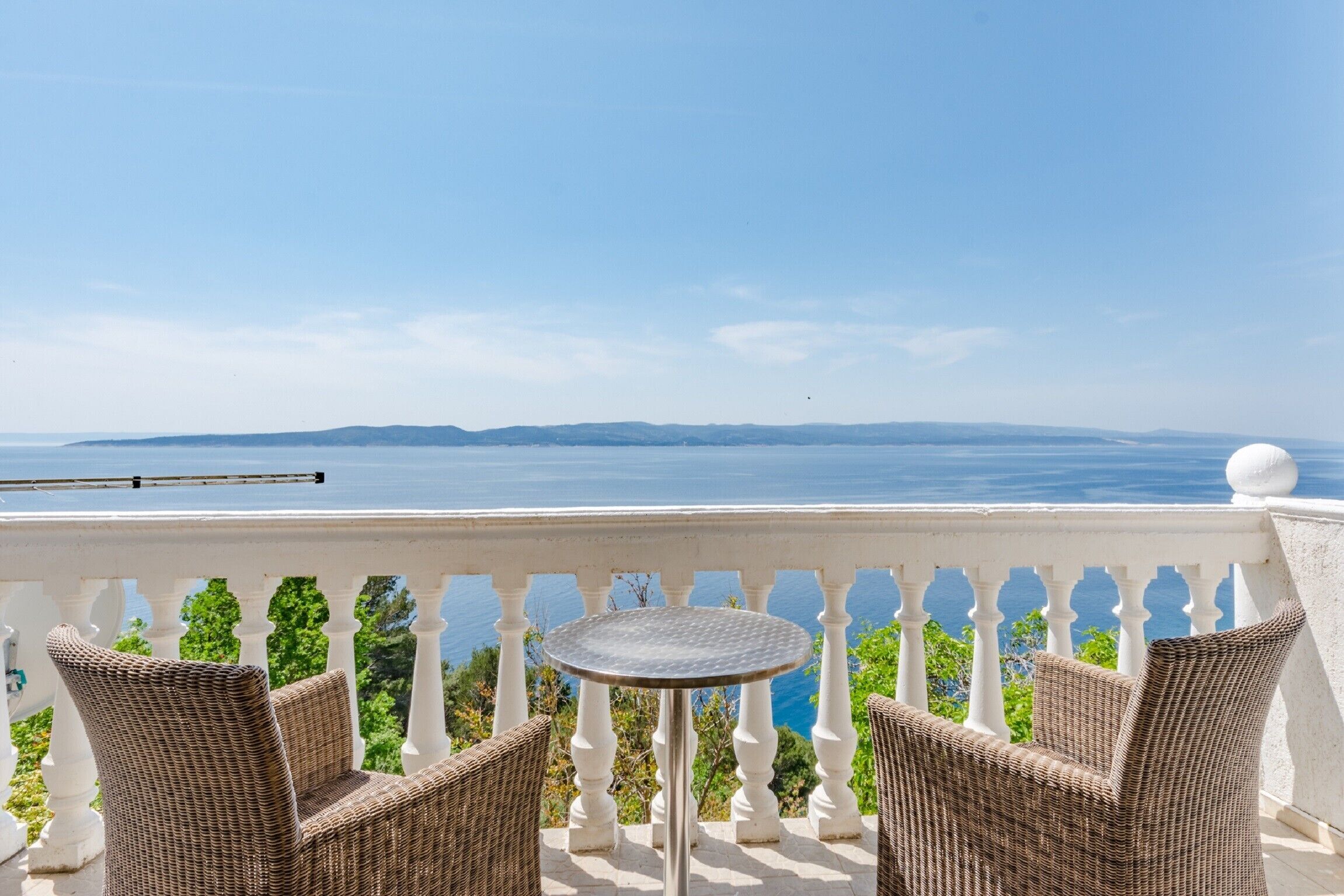 A balcony with wicker furniture and a small table, overlooking a scenic view of the sea and distant hills.