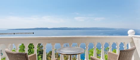 A balcony with wicker furniture and a small table, overlooking a scenic view of the sea and distant hills.