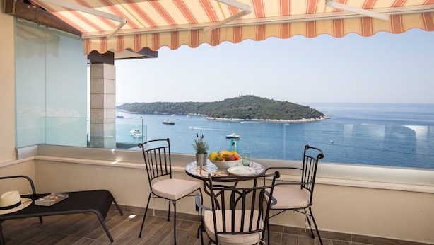 Ein Balkon mit gestreifter Markise, einem Tisch mit Stühlen, einem Blumentopf und Blick auf das Meer und eine ferne Insel.