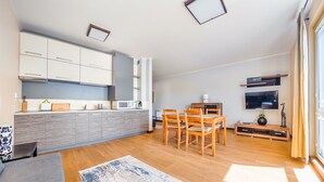 A modern kitchen with white cabinets, a dining area with wooden chairs, a TV mounted on the wall, and a large window with curtains. - Apartamenty Sun & Snow Niska (Krynica Morska)