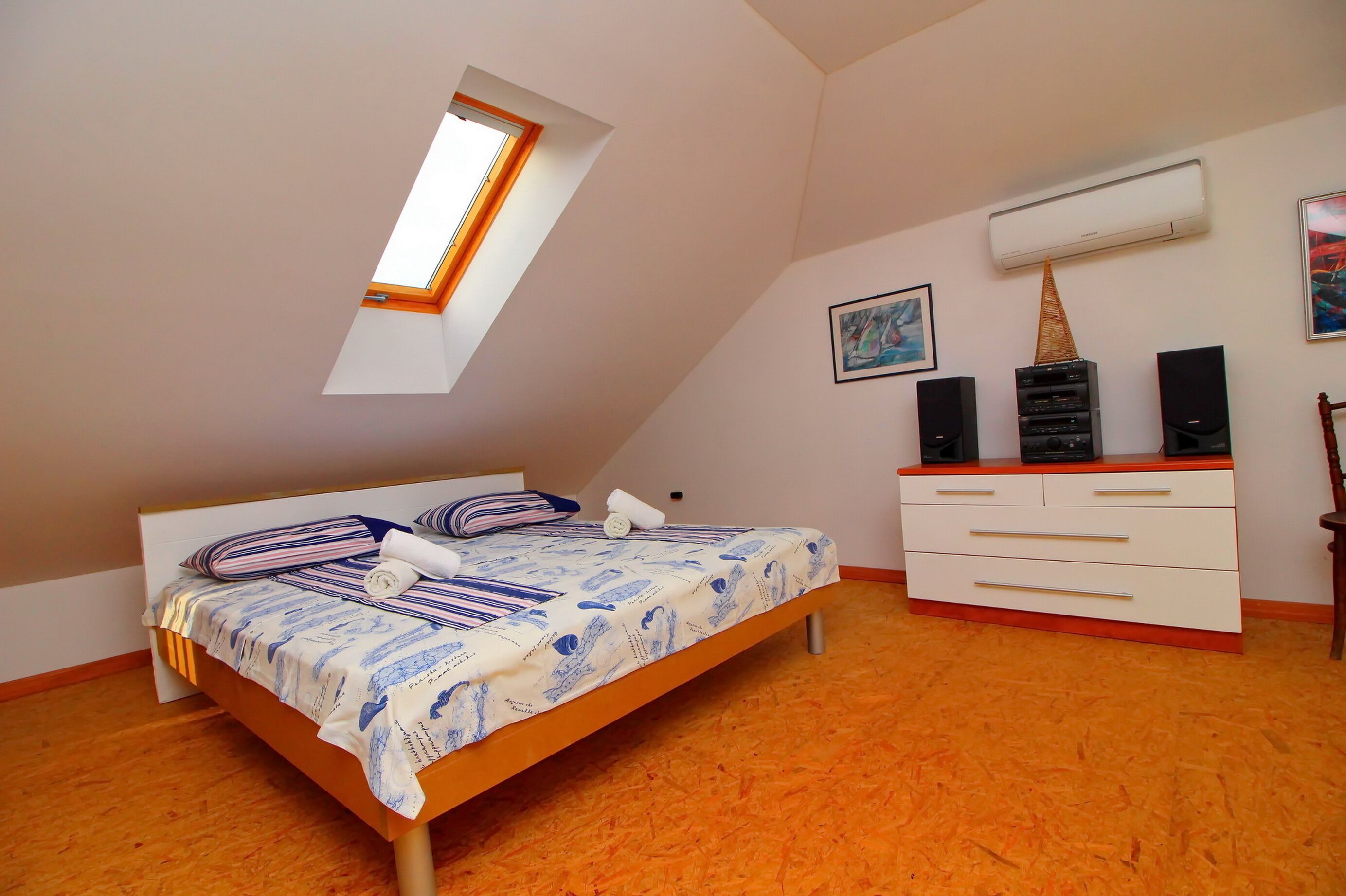 Un dormitorio con techo inclinado, tragaluz, cama con ropa de cama a rayas, cómoda blanca y dos altavoces en la pared.