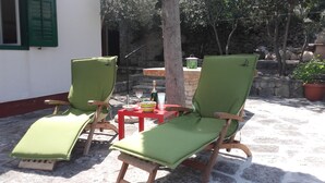 Twee groene lounge fauteuils met houten frames en kussens, een klein rood tafeltje met een fles en glazen, en een stenen muur op de achtergrond.