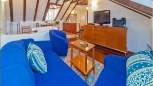 Ein Loft-Wohnzimmer mit freiliegenden Holzbalken, einem blauen Sofagarnitur, einem Holztisch und einem Fernseher auf einem Holzschrank.